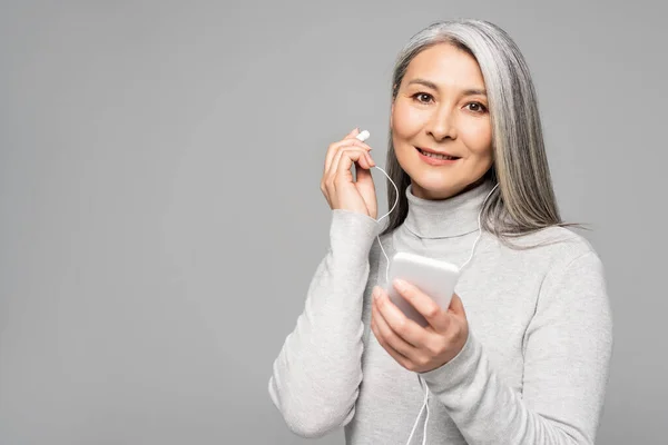 Happy asian woman in turtleneck with grey hair listening music with earphones and smartphone isolated on grey — Stock Photo