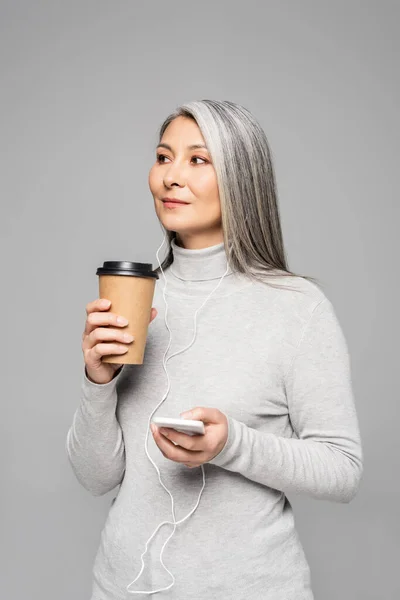 Beautiful asian woman in turtleneck with coffee to go listening music with earphones and smartphone isolated on grey — Stock Photo