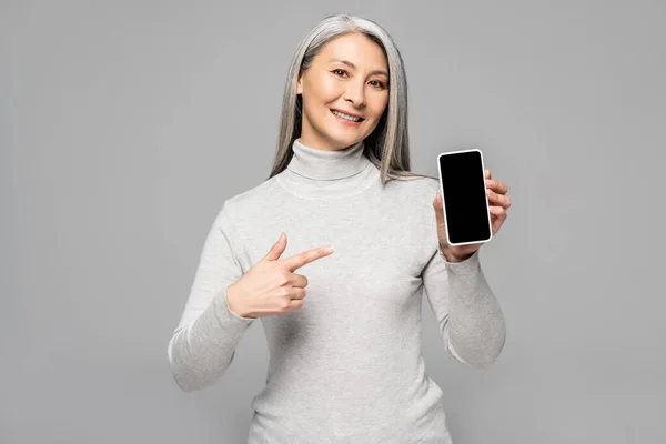 Mujer asiática feliz en cuello alto con pelo gris apuntando a teléfono inteligente con pantalla en blanco aislado en gris - foto de stock