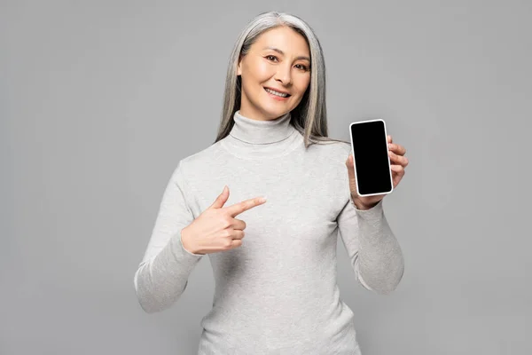 Happy asian woman in turtleneck with grey hair pointing at smartphone with blank screen isolated on grey — Stock Photo