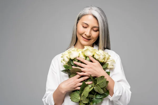 Dankbare asiatische Frau mit grauen Haaren, die einen Strauß weißer Rosen auf grau isoliert hält — Stockfoto