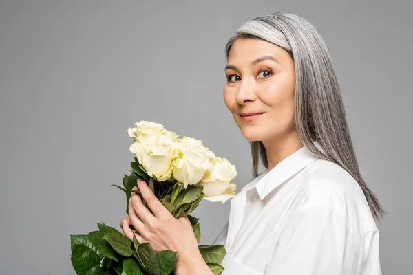 Adult smiling asian woman with grey hair holding bouquet of white roses isolated on grey — Stock Photo