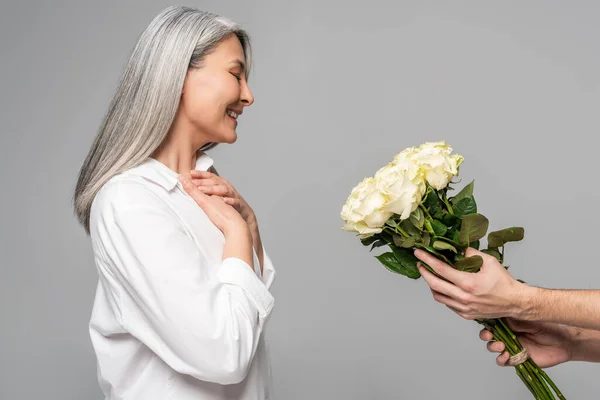 Feliz adulto asiático mulher com cabelos grisalhos em branco camisa olhando para buquê de rosas brancas de homem isolado no cinza — Fotografia de Stock