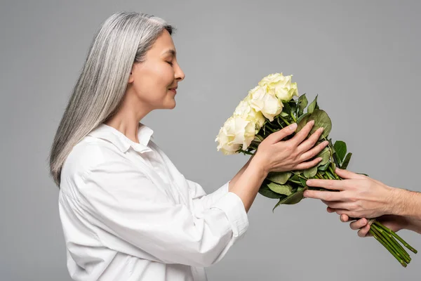 Positive adult asian woman with grey hair in white shirt taking bouquet of white roses from man isolated on grey — Stock Photo
