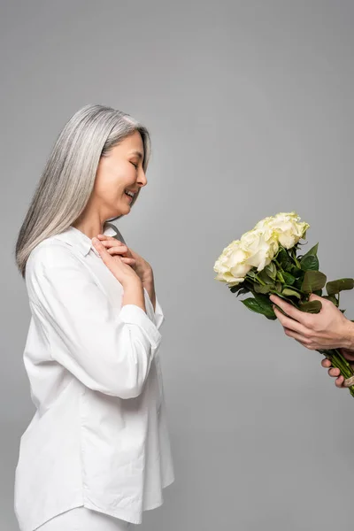Cheerful adult asian woman with grey hair in white shirt taking bouquet of white roses from man isolated on grey — Stock Photo