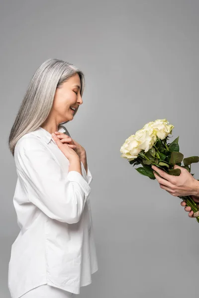 Alegre adulto asiático mulher com cinza cabelo no branco camisa tomando buquê de branco rosas de homem isolado no cinza — Fotografia de Stock