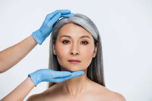 Médico en guantes de látex tocando la cara de atractiva mujer asiática desnuda aislada en gris - foto de stock