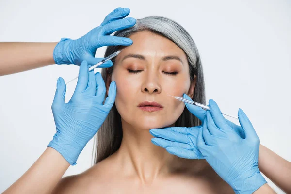 Mujer asiática desnuda y médicos en guantes de látex sosteniendo jeringas con inyecciones de belleza aisladas en gris - foto de stock