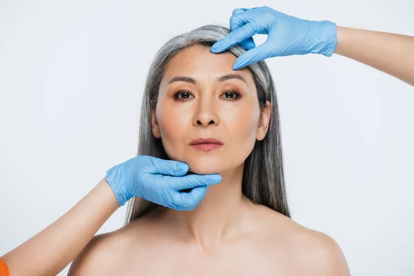 Médicos en guantes de látex tocando la cara de mujer asiática desnuda con el pelo gris aislado en gris - foto de stock