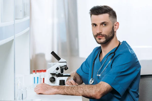 Biólogo joven serio mirando la cámara mientras está sentado cerca del microscopio - foto de stock