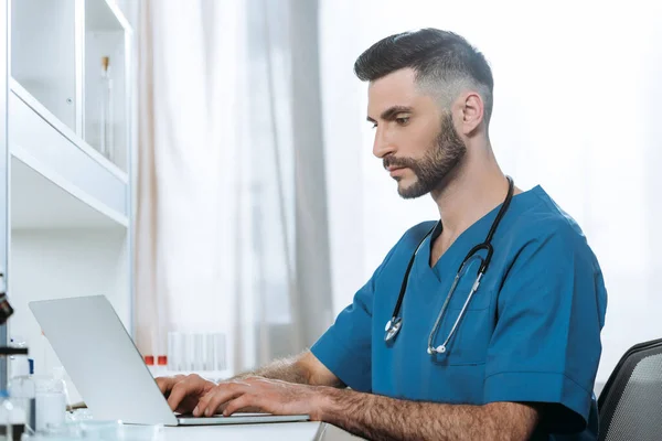 Médico joven con estetoscopio en el cuello escribiendo en el ordenador portátil - foto de stock