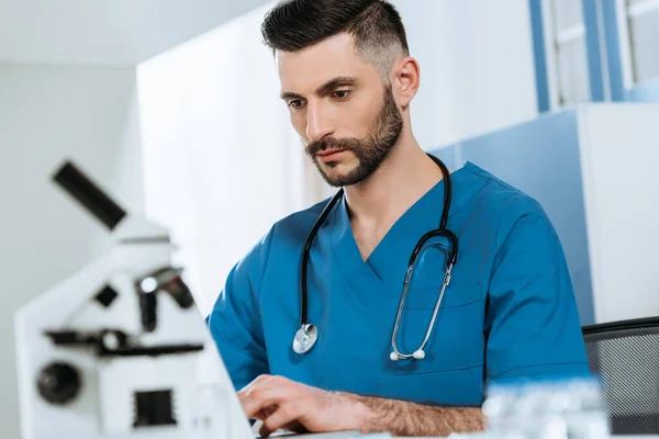 Selective focus of concentrated biologist typing on laptop near microscope — Stock Photo