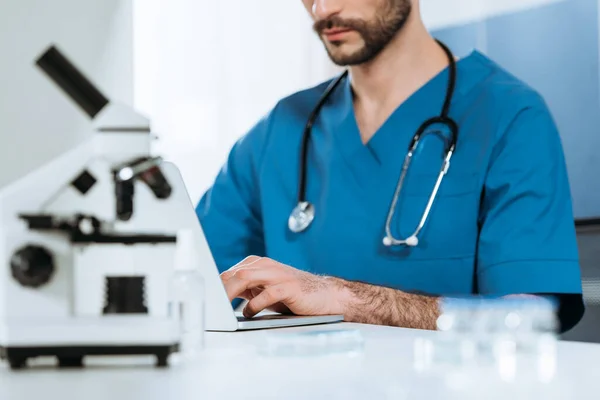 Vista recortada del joven biólogo escribiendo en el ordenador portátil cerca del microscopio - foto de stock