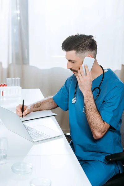 High angle view of young biologist writing while talking on smartphone near laptop and test tubes — Stock Photo