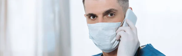 Fotografía panorámica del joven médico en máscara médica y guantes de látex hablando en smartphone y mirando a la cámara - foto de stock