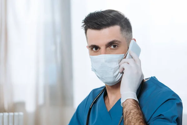 Молодой врач в медицинской маске и латексных перчатках разговаривает на смартфоне и смотрит в камеру — стоковое фото