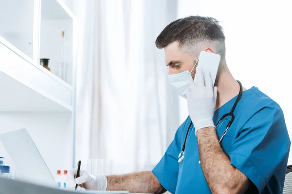 Селективное внимание молодого врача в медицинской маске и латексных перчатках, пишущего во время разговора на смартфоне — стоковое фото