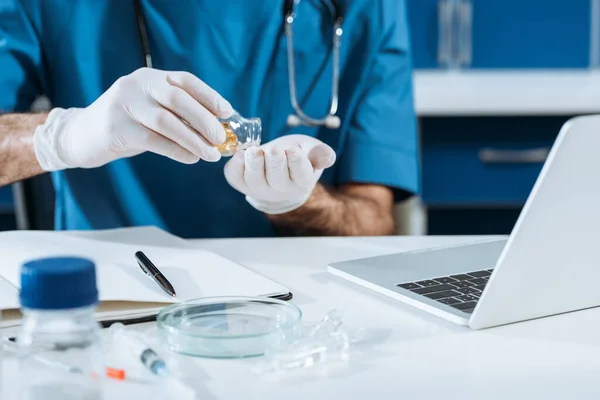 Cropped view of scientist in latex gloves taking capsules from glass container near laptop — Stock Photo