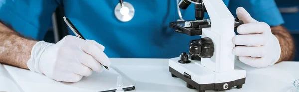 Vista parcial del biólogo en guantes de látex escritura en cuaderno cerca del microscopio, plano panorámico - foto de stock