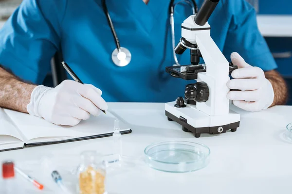 Vista recortada del biólogo en guantes de látex escritura en cuaderno cerca del microscopio - foto de stock