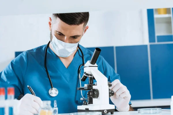 Selective focus of young biologist in medical mask and latex gloves writing near microscope in lab — Stock Photo