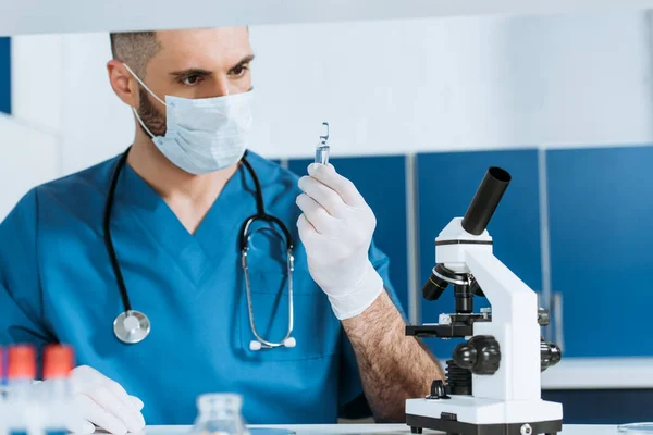 Селективный фокус биолога в медицинской маске и латексных перчатках, держащих ампулу с лекарством рядом с микроскопом — стоковое фото