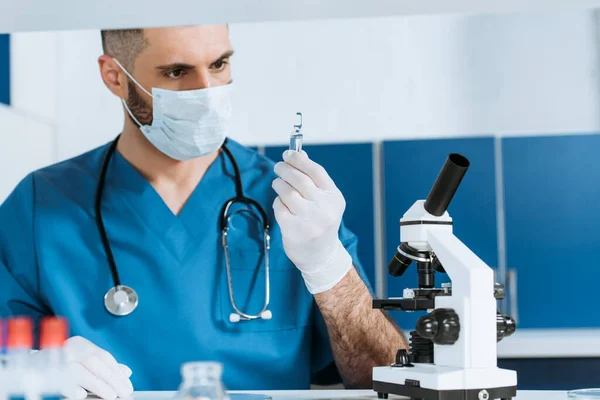 Foyer sélectif du biologiste dans le masque médical et les gants en latex tenant ampule avec la médecine près du microscope — Photo de stock