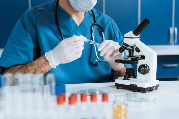 Cropped view of biologist in medical mask and latex gloves holding syringe near microscope — Stock Photo