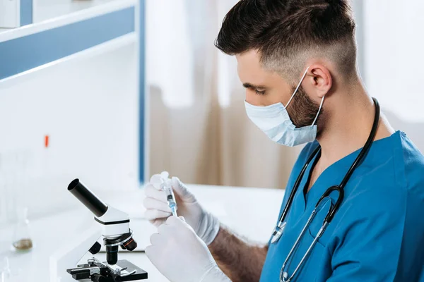 Jeune biologiste en masque médical et gants en latex tenant la seringue près du microscope — Photo de stock