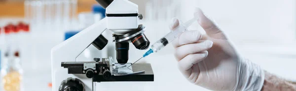 Частковий вид біолога в латексних рукавичках, що тримає шприц біля мікроскопа, панорамний знімок — стокове фото