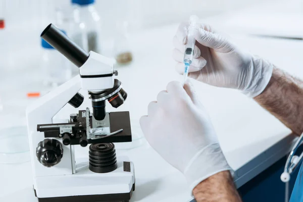 Частичный обзор биолога в латексных перчатках, держащего шприц рядом с микроскопом — стоковое фото