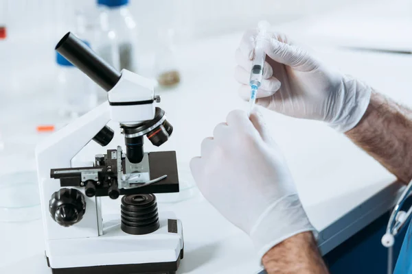 Vista parcial del biólogo en guantes de látex que sostienen la jeringa cerca del microscopio - foto de stock