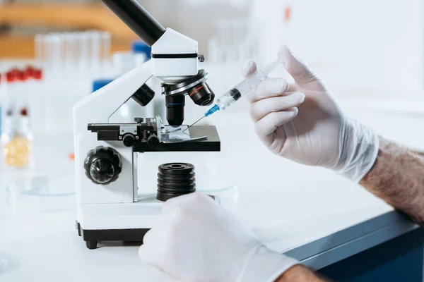 Vista recortada del biólogo en guantes de látex que sostienen la jeringa cerca del microscopio - foto de stock