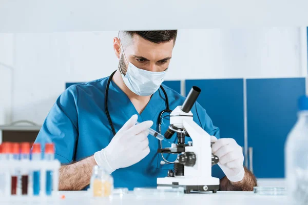 Enfoque selectivo del biólogo en máscara médica y guantes de látex que sostienen la jeringa cerca del microscopio - foto de stock