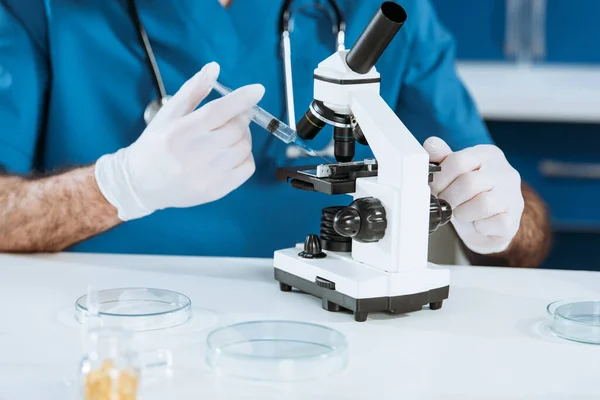 Ausgeschnittene Ansicht eines Biologen in Latexhandschuhen, der Spritze in der Nähe des Mikroskops hält — Stockfoto