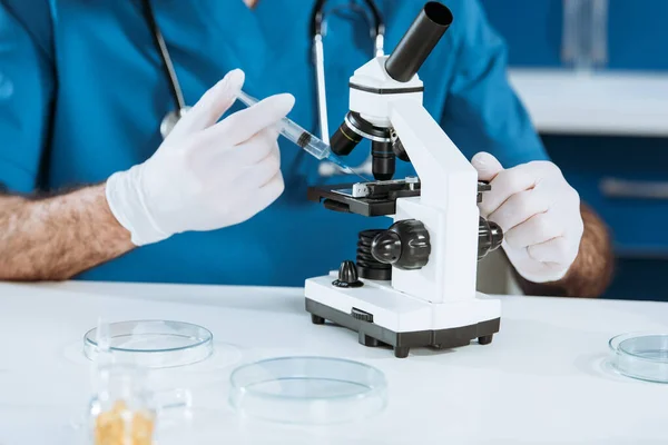 Vista recortada del biólogo en guantes de látex que sostienen la jeringa cerca del microscopio - foto de stock