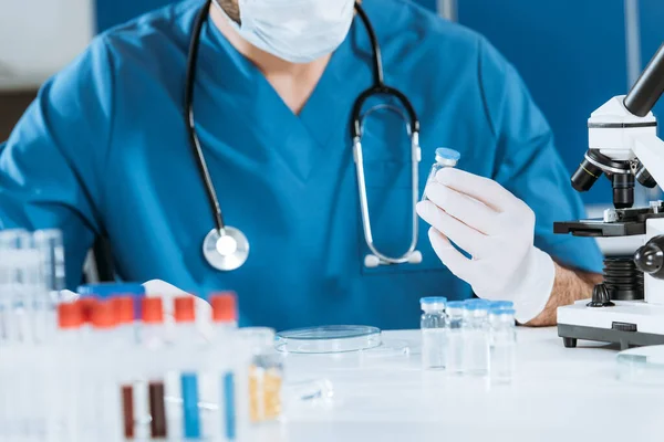 Cropped view of biologist in latex gloves holding glass container with vaccine near test tubes — Stock Photo