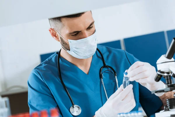 Молодой врач в медицинской маске и латексных перчатках, держащий стеклянный контейнер с лекарствами — стоковое фото