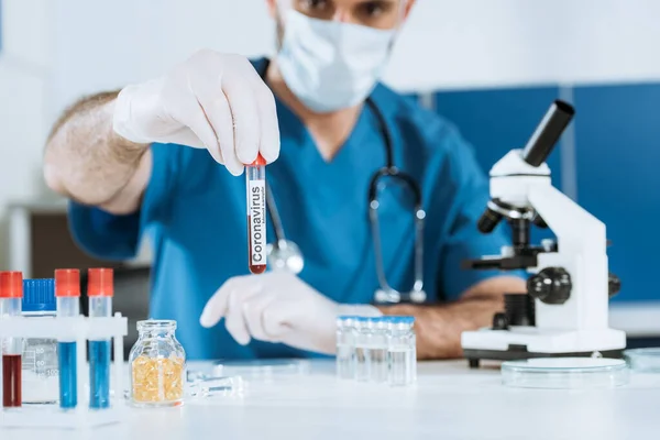 Enfoque selectivo del científico en máscara médica y guantes de látex que sostienen el tubo de ensayo con inscripción de coronavirus - foto de stock