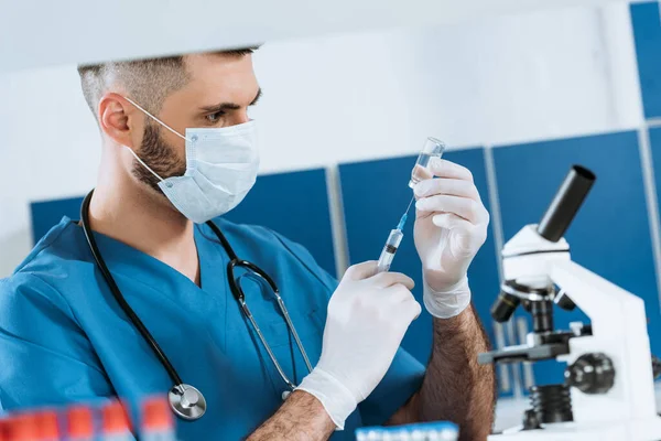 Médico joven con máscara médica y guantes de látex que toma medicamentos con jeringa - foto de stock