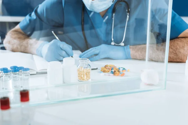 Vista cortada do veterinário em máscara médica e luvas tardias escrevendo perto de recipientes com medicamentos e mouse branco em caixa de vidro — Fotografia de Stock