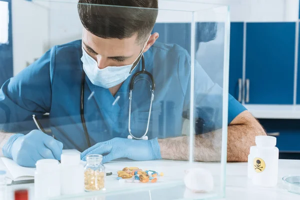 Jovem veterinário em máscara médica e luvas atrasadas escrevendo perto de recipientes com medicamentos e mouse branco em caixa de vidro — Fotografia de Stock