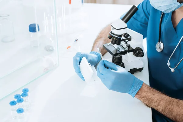 Обрізаний вид біолога в медичній масці та рукавичках, що тримають пробну коробку під час аналізу мікроскопом біля білої миші в скляній коробці — стокове фото
