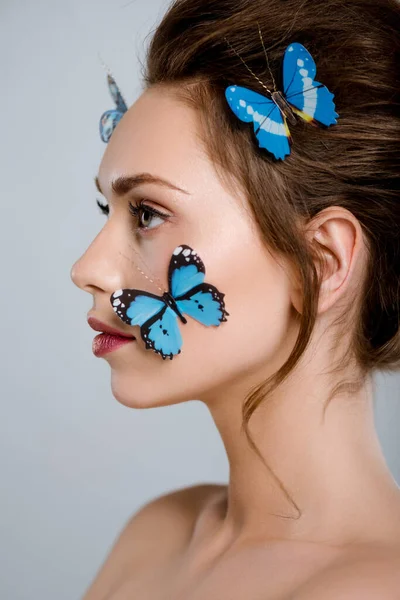 Привлекательная женщина с голубыми декоративными бабочками на лице, изолированными от серого — стоковое фото