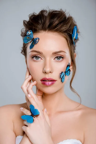 Mulher atraente com borboletas decorativas azuis no rosto olhando para a câmera isolada no cinza — Fotografia de Stock
