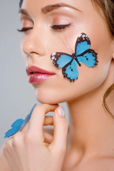 Привлекательная женщина с закрытыми глазами и голубыми декоративными бабочками на лице, изолированными на сером — стоковое фото