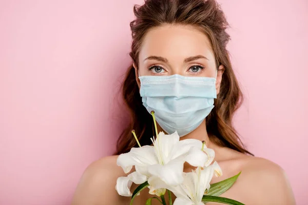 Молода жінка в медичній масці дивиться на камеру і тримає квіти ізольовані на рожевому — стокове фото