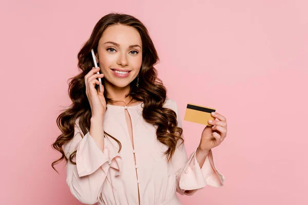 Mujer joven y feliz hablando en el teléfono inteligente y la celebración de la tarjeta de crédito aislado en rosa - foto de stock