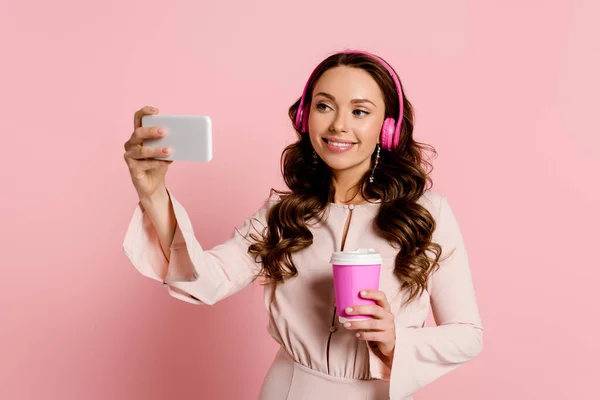 Chica feliz en auriculares inalámbricos tomando selfie y sosteniendo taza de papel en rosa - foto de stock