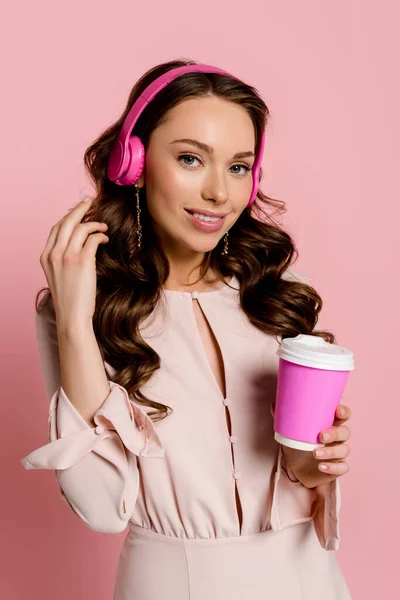 Alegre chica escuchando música en auriculares inalámbricos y sosteniendo taza de papel aislado en rosa - foto de stock